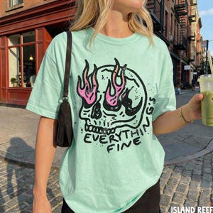 May include: A light mint green short-sleeve t-shirt featuring a black graphic of a skull with pink flames in the eye sockets and the text "EVERYTHING'S FINE" printed below. The shirt is paired with a black shoulder bag with a thin strap.