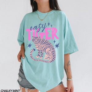 Puede incluir: Una camiseta azul claro con las palabras "easy TIGER" en rosa. La camiseta presenta un gr&aacute;fico de tigre rosa y azul y acentos de estrellas. Las palabras "CHALKY MINT" est&aacute;n en la parte inferior.