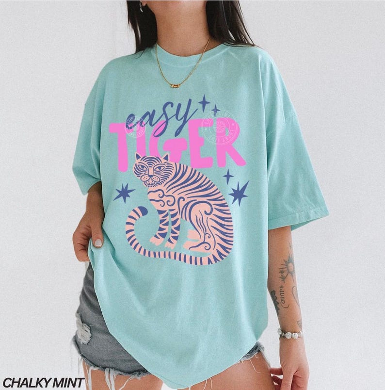 Puede incluir: Una camiseta azul claro con las palabras "easy TIGER" en rosa. La camiseta presenta un gr&aacute;fico de tigre rosa y azul y acentos de estrellas. Las palabras "CHALKY MINT" est&aacute;n en la parte inferior.