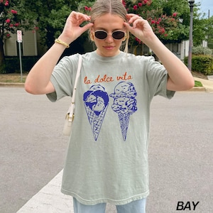 May include: A light green t-shirt featuring a graphic of two ice cream cones. The text "la dolce vita" is printed in orange above the ice cream cones.