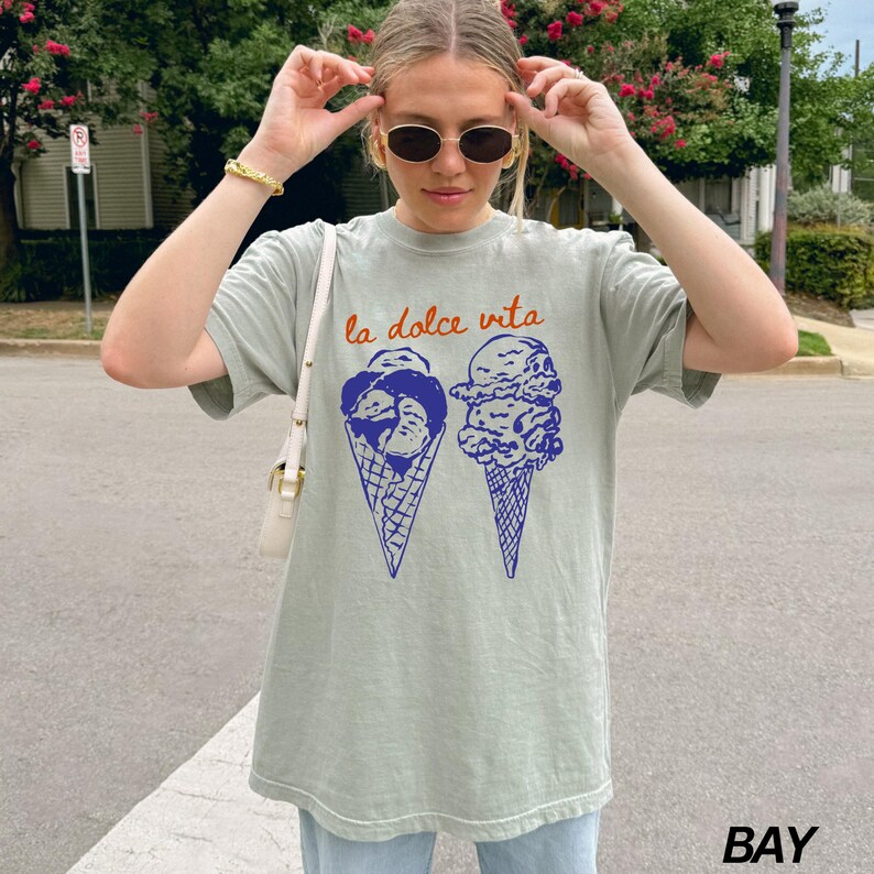 May include: A light green t-shirt featuring a graphic of two ice cream cones. The text "la dolce vita" is printed in orange above the ice cream cones.