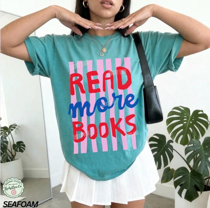Puede incluir: Camiseta verde mar con las palabras "READ more BOOKS" en rojo, azul y rosa. El texto est&aacute; rayado verticalmente con rosa. La camiseta tiene mangas cortas y un corte holgado. Parte del dise&ntilde;o original de "Lucky Lime Co".