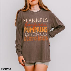 May include: A long-sleeve espresso brown t-shirt featuring the text FLANNELS, bonfires, PUMPKINS, football, and HAYRIDES printed in a mix of cream, orange, and rust colors on the front. The shirt has a crew neckline and a relaxed fit.