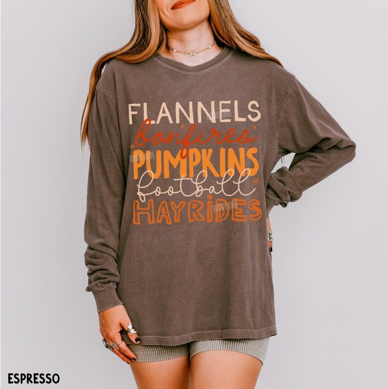 May include: A long-sleeve espresso brown t-shirt featuring the text FLANNELS, bonfires, PUMPKINS, football, and HAYRIDES printed in a mix of cream, orange, and rust colors on the front. The shirt has a crew neckline and a relaxed fit.