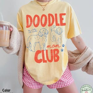 Puede incluir: Una camiseta amarilla con las palabras "DOODLE MOM CLUB" en letras rojas. La camiseta presenta dos ilustraciones de perros doodle y la frase "life is better with doodles". La camiseta es de color pl&aacute;tano.