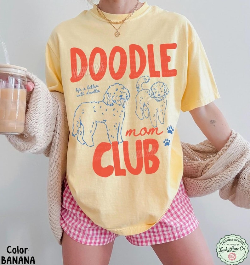 Puede incluir: Una camiseta amarilla con las palabras "DOODLE MOM CLUB" en letras rojas. La camiseta presenta dos ilustraciones de perros doodle y la frase "life is better with doodles". La camiseta es de color pl&aacute;tano.