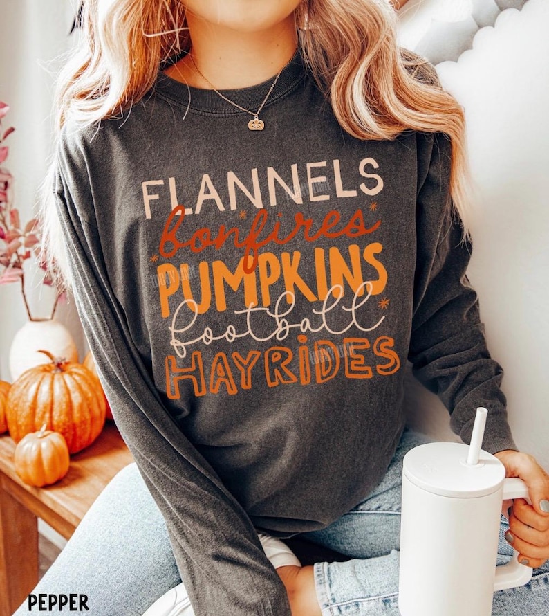 May include: A charcoal gray long-sleeve crewneck sweatshirt featuring the text FLANNELS, bonfires, PUMPKINS, football, and HAYRIDES in various orange, cream, and brown fonts. The shirt has a relaxed fit and a vintage-washed appearance.