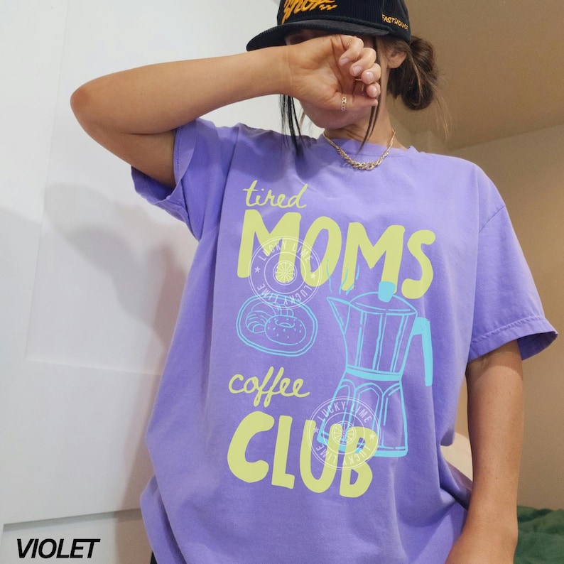 May include: Violet short-sleeve t-shirt with the text "tired MOMS coffee CLUB" in yellow and light blue. The graphic includes a coffee pot and a pastry. The word "VIOLET" is printed at the bottom left.