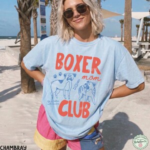 May include: A light blue short-sleeve t-shirt featuring the red text BOXER CLUB and the blue text mom. The graphic includes two line-art illustrations of boxer dogs, paw prints, and the small blue text life is better with boxers.