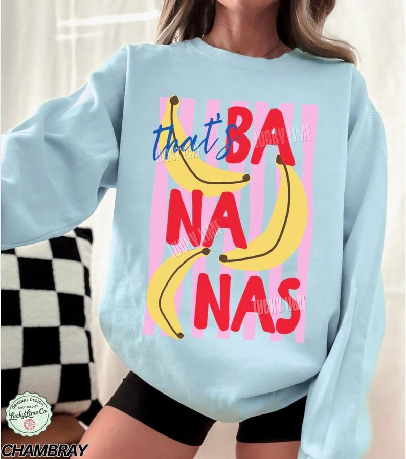 May include: Light blue sweatshirt with a graphic design featuring the words "that's BANANAS" in a playful font, with yellow banana illustrations and pink vertical stripes. The sweatshirt has long sleeves.