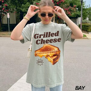 May include: A light green t-shirt with a graphic of a grilled cheese sandwich and the text "Grilled Cheese" in brown letters. The sandwich has extra cheese and the text "Extra Cheesy" is in a starburst shape.