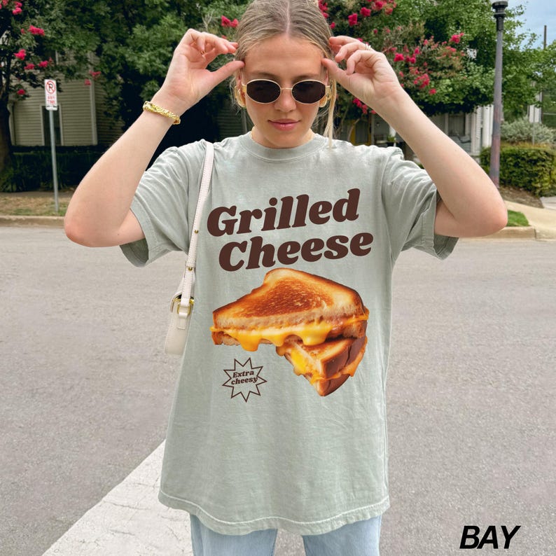 May include: A light green t-shirt with a graphic of a grilled cheese sandwich and the text "Grilled Cheese" in brown letters. The sandwich has extra cheese and the text "Extra Cheesy" is in a starburst shape.