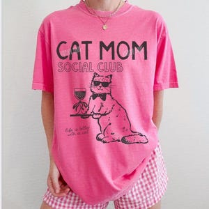 K&ouml;nnte beinhalten: Ein leuchtend pinkes Kurzarm-T-Shirt mit schwarzem Text, der CAT MOM SOCIAL CLUB und Life is better with a cat lautet. Die Grafik zeigt eine Katze mit Sonnenbrille und Fliege, die ein Weinglas auf einem Tablett h&auml;lt, gedruckt in schwarzer Tinte.