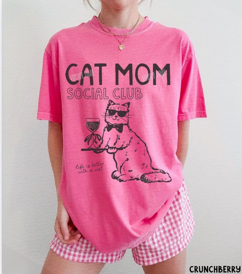 K&ouml;nnte beinhalten: Ein leuchtend pinkes Kurzarm-T-Shirt mit schwarzem Text, der CAT MOM SOCIAL CLUB und Life is better with a cat lautet. Die Grafik zeigt eine Katze mit Sonnenbrille und Fliege, die ein Weinglas auf einem Tablett h&auml;lt, gedruckt in schwarzer Tinte.