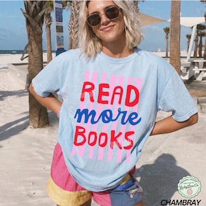 Puede incluir: Camiseta azul claro con las palabras "READ more BOOKS" en rojo y azul. El texto est&aacute; sobre un fondo de rayas rosas. La camiseta es de la colecci&oacute;n "Chambray".