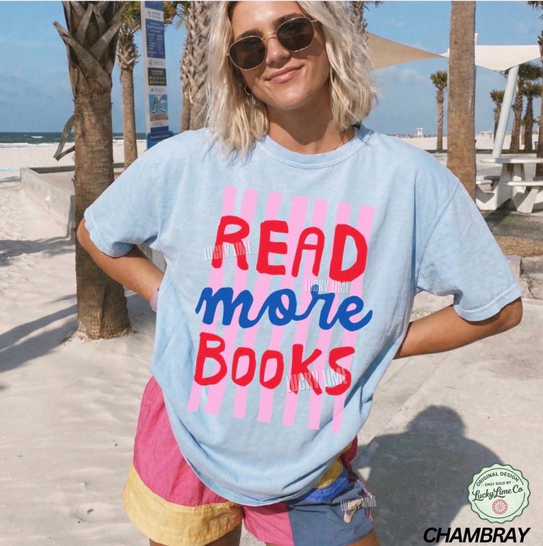 Puede incluir: Camiseta azul claro con las palabras "READ more BOOKS" en rojo y azul. El texto est&aacute; sobre un fondo de rayas rosas. La camiseta es de la colecci&oacute;n "Chambray".