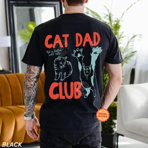 Peut inclure: T-shirt noir avec l'inscription "CAT DAD CLUB" en orange, orn&eacute; d'illustrations de chats en blanc et turquoise. L'expression "life is better with cats" est &eacute;galement imprim&eacute;e. Le t-shirt a un aspect vintage et us&eacute;.