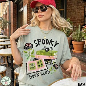 Op de afbeelding: Saliegroen t-shirt met de tekst "Spooky Book Club" en een afbeelding van een spin op boeken, rozen en een open boek. Het shirt heeft een vintage, licht vervaagde uitstraling.