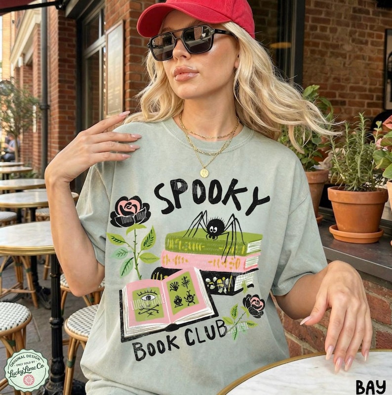 Op de afbeelding: Saliegroen t-shirt met de tekst "Spooky Book Club" en een afbeelding van een spin op boeken, rozen en een open boek. Het shirt heeft een vintage, licht vervaagde uitstraling.