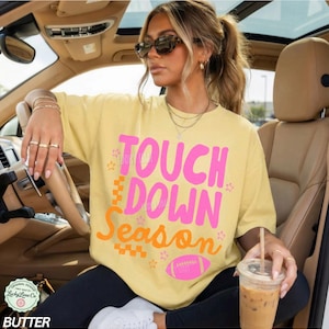 May include: Butter yellow t-shirt with the text "Touch Down Season" in pink and orange, featuring a football graphic. The shirt has a relaxed fit and is worn by a person in a car.
