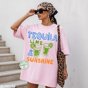 May include: A light pink oversized t-shirt with the words "TEQUILA LIME & SUNSHINE" in blue, green, and orange. The shirt features a graphic of two cocktails with lime wedges. A leopard print tote bag is visible.