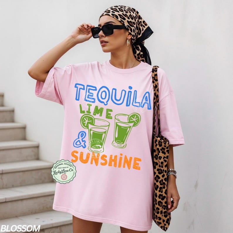 May include: A light pink oversized t-shirt with the words "TEQUILA LIME & SUNSHINE" in blue, green, and orange. The shirt features a graphic of two cocktails with lime wedges. A leopard print tote bag is visible.
