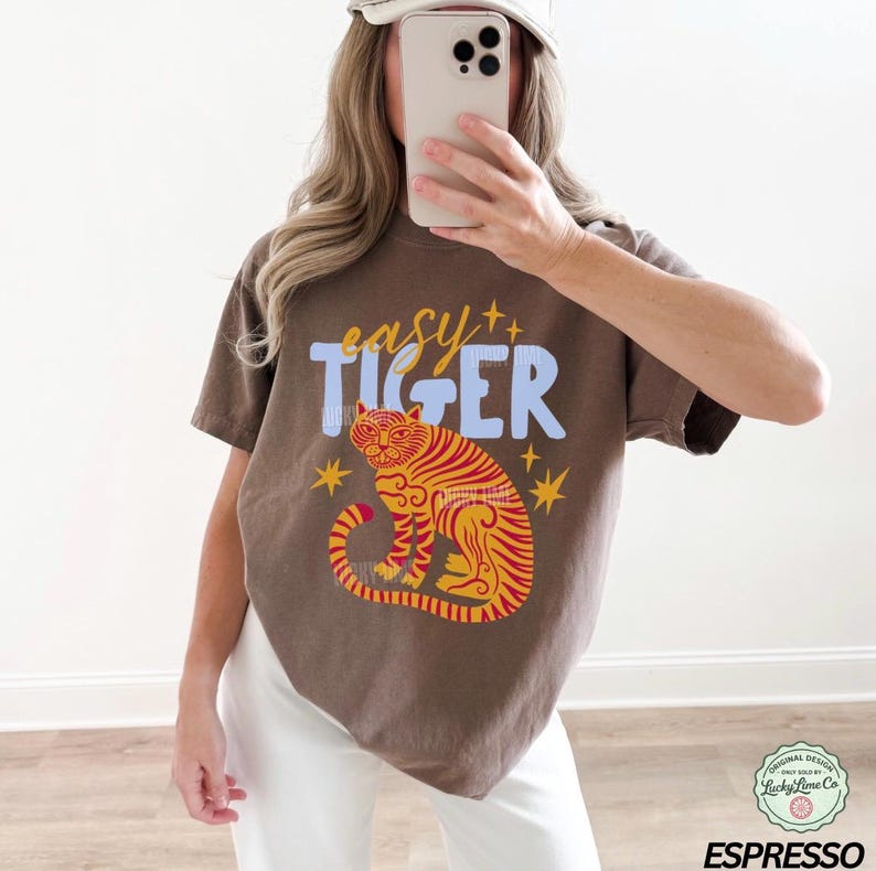 May include: Espresso-colored t-shirt featuring the text "easy TIGER" above a stylized orange and red tiger illustration. The design includes star accents. The shirt is a casual style.