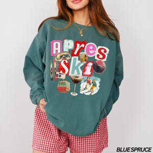 May include: Teal-colored sweatshirt with the words "Apres Ski" in a collage style. The design includes ski goggles, cocktails, a disco ball, a ski lift, and skiers. The text "BLUE SPRUCE" is printed at the bottom right.