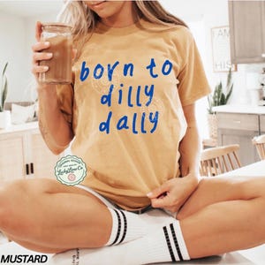 May include: Mustard-colored t-shirt with the phrase "born to dilly dally" in blue script. The shirt features a round logo at the bottom and is paired with white shorts and striped socks. A glass of iced coffee is held in the hand.