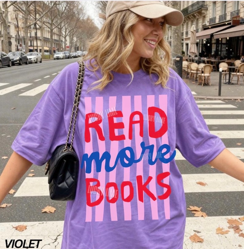 Puede incluir: Camiseta violeta con la frase "READ more BOOKS" en letras rojas y azules. El texto se encuentra sobre un fondo a rayas rosas y moradas. La camiseta es de estilo casual y holgado.