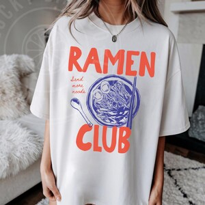 May include: White oversized t-shirt with a graphic of a bowl of ramen noodles and the text "Ramen Club" with the words "send more noodles" underneath.