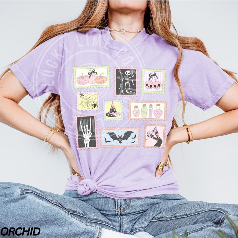 May include: A light purple t-shirt featuring a collection of Halloween-themed postage stamp designs. The stamps depict pumpkins, a skeleton, cherries, a spider web, a witch's hat, potion bottles, a skeleton hand, bats, and a cat.