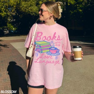 以下が含まれることがあります： 「Books are my Love Language」の文字が書かれたピンク色のTシャツ。本、ティーカップ、ハートのイラストが描かれています。ゆったりとしたオーバーサイズのカジュアルなデザインです。