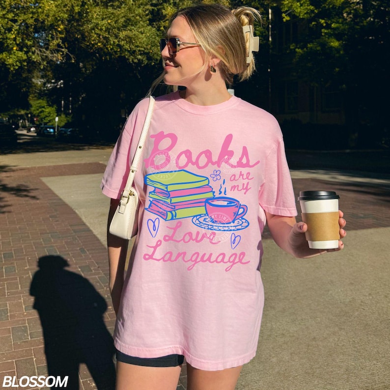 以下が含まれることがあります： 「Books are my Love Language」の文字が書かれたピンク色のTシャツ。本、ティーカップ、ハートのイラストが描かれています。ゆったりとしたオーバーサイズのカジュアルなデザインです。