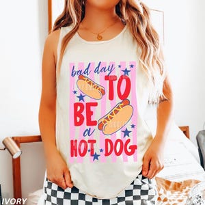 Op de afbeelding: Ivoor tanktop met de tekst "bad day to be a hot dog" in roze en rood, met hotdog- en sterrenafbeeldingen. De tekst staat in het midden op de voorkant van de mouwloze top.