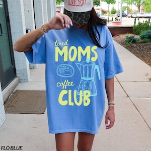 May include: A light blue t-shirt with the text "tired MOMS coffee CLUB" in yellow and light blue. The shirt features a coffee pot graphic and a pastry graphic. The shirt is a Flo Blue color.