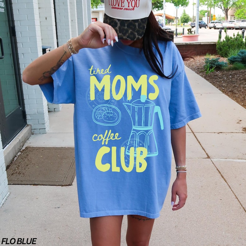 May include: A light blue t-shirt with the text "tired MOMS coffee CLUB" in yellow and light blue. The shirt features a coffee pot graphic and a pastry graphic. The shirt is a Flo Blue color.