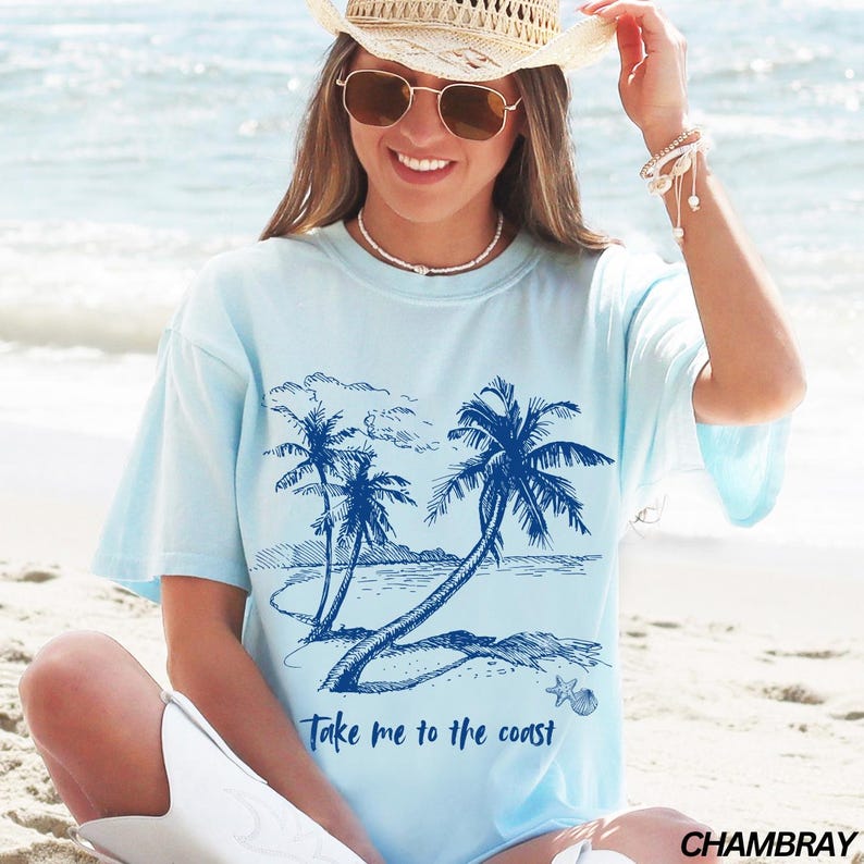 May include: A light blue t-shirt with a tie-dye effect featuring a blue graphic of palm trees on a beach with the text "Take me to the coast". The shirt is made of a soft, comfortable chambray fabric.