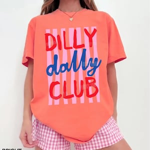 May include: A coral-colored t-shirt with the words "DILLY dally CLUB" in red and blue, with pink vertical stripes. The model is wearing pink and white checkered shorts. The words "BRIGHT SALMON" are at the bottom left.