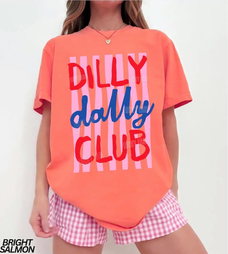 May include: A coral-colored t-shirt with the words "DILLY dally CLUB" in red and blue, with pink vertical stripes. The model is wearing pink and white checkered shorts. The words "BRIGHT SALMON" are at the bottom left.