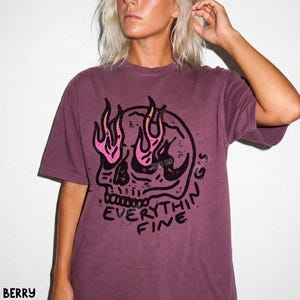 May include: A berry-colored short-sleeve t-shirt featuring a black graphic of a skull with pink and orange flames rising from the eye sockets. Below the skull, the text reads EVERYTHING FINE in a bold, distressed black font style.