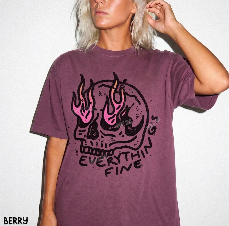 May include: A berry-colored short-sleeve t-shirt featuring a black graphic of a skull with pink and orange flames rising from the eye sockets. Below the skull, the text reads EVERYTHING FINE in a bold, distressed black font style.