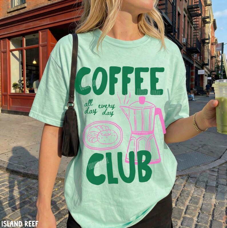 Peut inclure: Un t-shirt vert clair avec le texte vert fonc&eacute; COFFEE CLUB et la phrase all day day. Le v&ecirc;tement pr&eacute;sente un dessin au trait rose repr&eacute;sentant une cafeti&egrave;re italienne et une p&acirc;tisserie sur une assiette. Un sac &agrave; bandouli&egrave;re noir est port&eacute;.