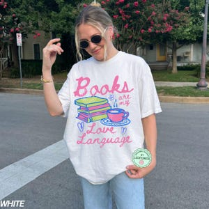 以下が含まれることがあります： ピンクと青で「Books are my Love Language」と書かれた白いTシャツ。グラフィックには、本の山、ティーカップ、ハートが含まれています。 ゆったりとしたオーバーサイズのフィット感のシャツです。