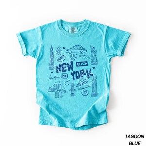 May include: Lagoon blue t-shirt with a New York City themed graphic. The design includes illustrations of the Statue of Liberty, the Empire State Building, a car, a pretzel, and the Brooklyn Bridge. The text "NEW YORK" is prominently displayed.