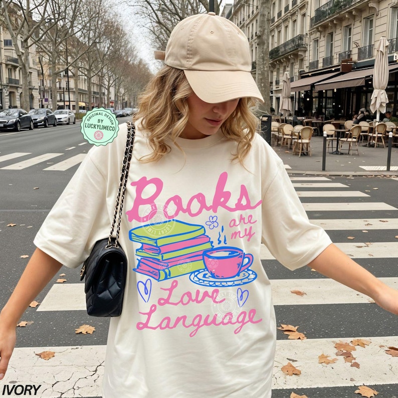 以下が含まれることがあります： 「Books are my Love Language」のピンクと青の文字がプリントされたアイボリー色のTシャツ。本の山、ティーカップ、ハートのグラフィック。ベージュのベースボールキャップと黒いハンドバッグがコーディネートを完成させています。
