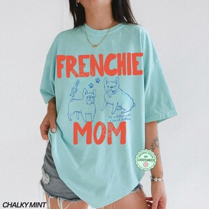 May include: A light blue t-shirt with the words "FRENCHIE MOM" in orange block letters. The shirt features blue line drawings of French Bulldogs and paw prints. The text "It's better with french bulldogs" is also on the shirt.