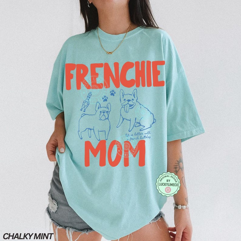 May include: A light blue t-shirt with the words "FRENCHIE MOM" in orange block letters. The shirt features blue line drawings of French Bulldogs and paw prints. The text "It's better with french bulldogs" is also on the shirt.