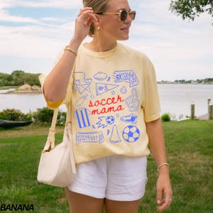 May include: A light yellow t-shirt with a blue and red graphic that says "soccer mama". The shirt has a crew neck and short sleeves. The person is wearing white shorts and sunglasses.