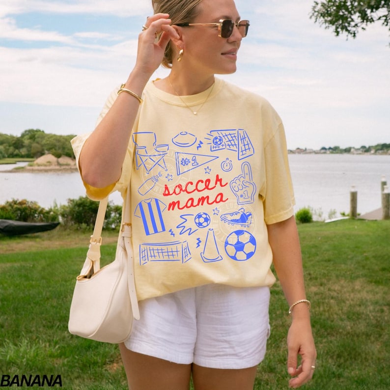 May include: A light yellow t-shirt with a blue and red graphic that says "soccer mama". The shirt has a crew neck and short sleeves. The person is wearing white shorts and sunglasses.